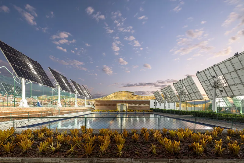 Reflecting pool with planted bed in foreground and solar panel canopies flanking a central pavilion at dusk