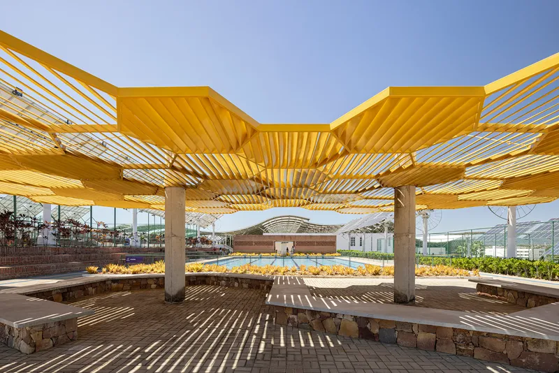 Seating area beneath the yellow slatted pergola with concrete columns and stone bench walls