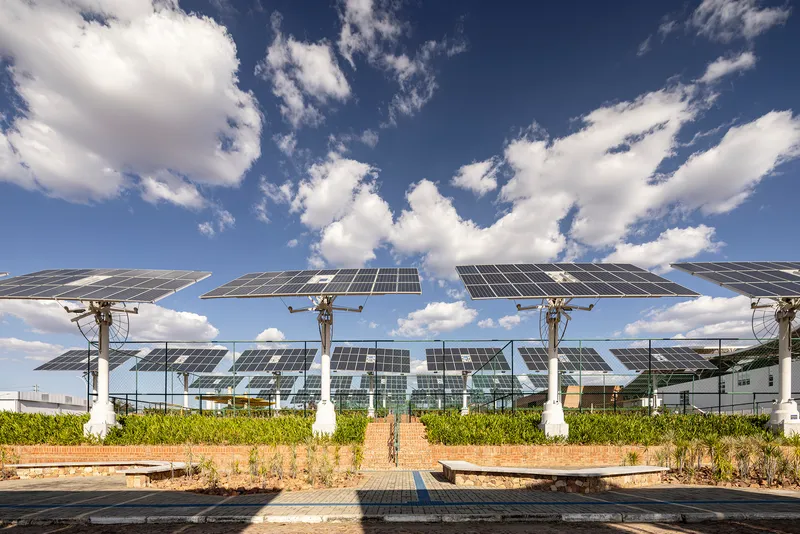 Row of tilted solar panels on white columns with pathway and scattered clouds overhead