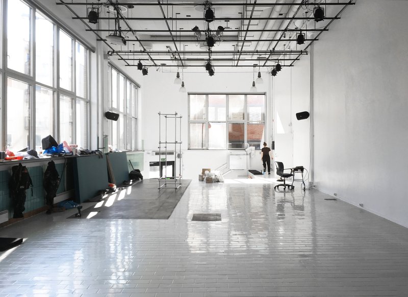 Open studio space with exposed ceiling grid, white tile floor and tall windows flooding the room with daylight