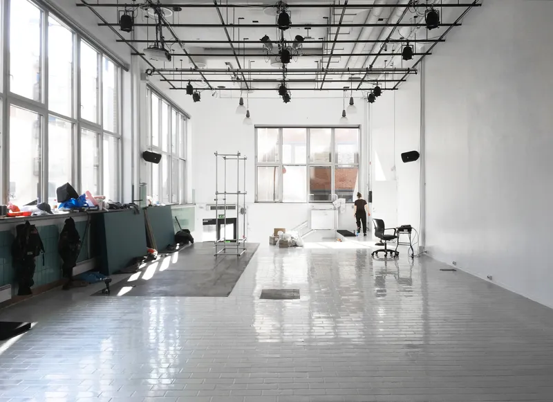 Open studio space with exposed ceiling grid, white tile floor and tall windows flooding the room with daylight
