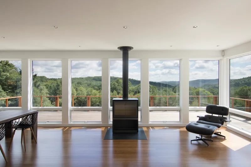 Open living space with freestanding black stove and full-height glazing facing forested hills