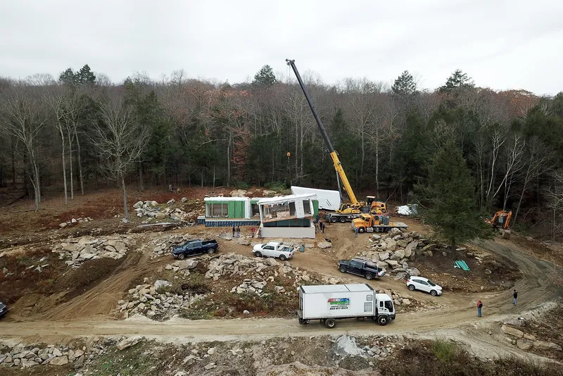Construction site with crane lifting prefabricated modules onto cleared hillside among bare winter trees