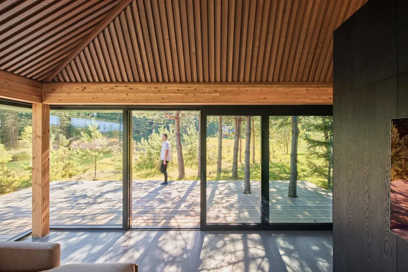 Interior view through floor-to-ceiling glazing to exterior terrace with timber slat ceiling above and figure standing outside