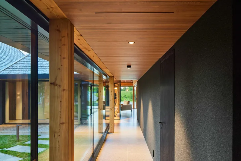 Glazed hallway with timber posts and plank ceiling looking toward another pavilion at dusk