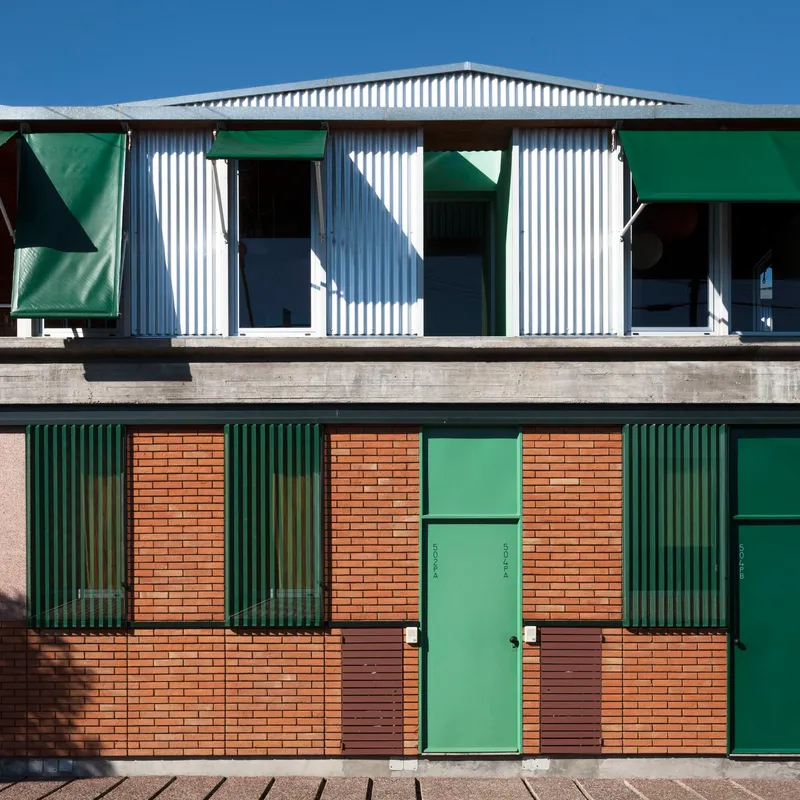 Two-storey facade with green corrugated metal shutters, orange brick panels, and white corrugated metal upper level under clear sky
