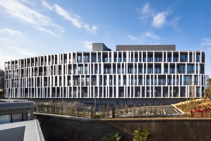 Street facade with vertical white and dark brise-soleil pattern rising above a concrete retaining wall with young trees