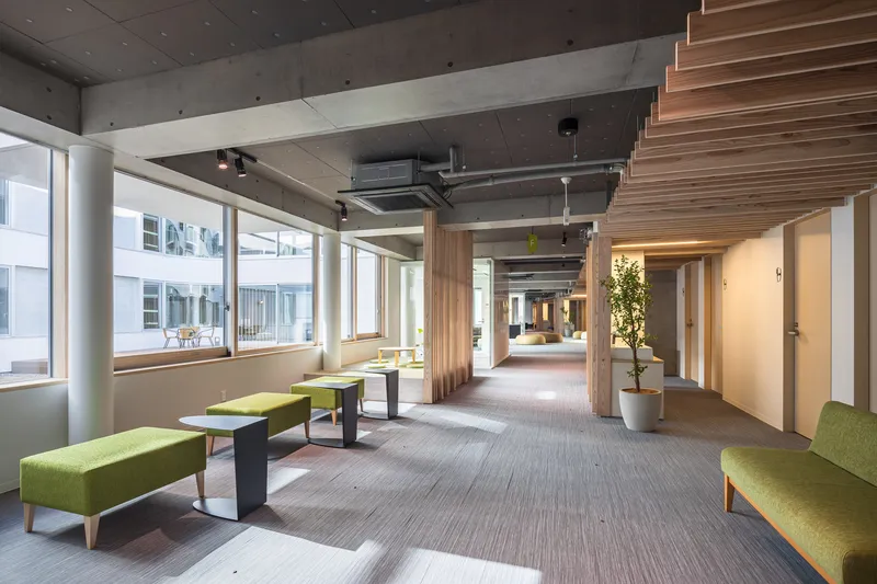 Interior corridor with exposed concrete ceiling, timber columns and green upholstered seating along windows overlooking a courtyard
