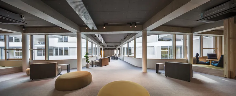 Open-plan common area with timber posts supporting concrete beams and continuous windows framing a neighboring facade