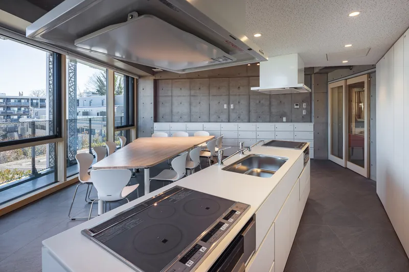 Shared kitchen with white island, integrated cooktop and concrete wall behind a dining table near corner windows