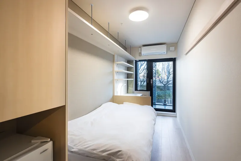 Compact bedroom with built-in plywood storage, suspended shelving and window view to bare winter trees outside