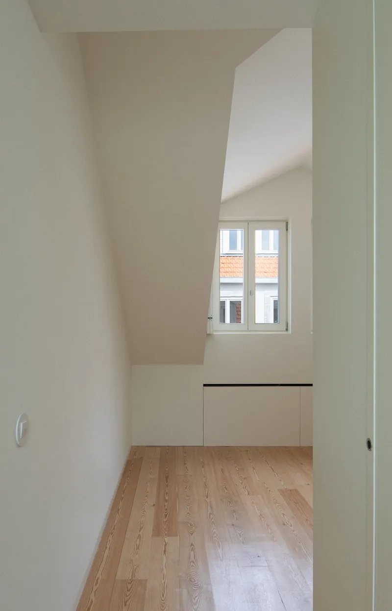 Small bedroom with oak flooring and low built-in cabinet below a window with rooftop view