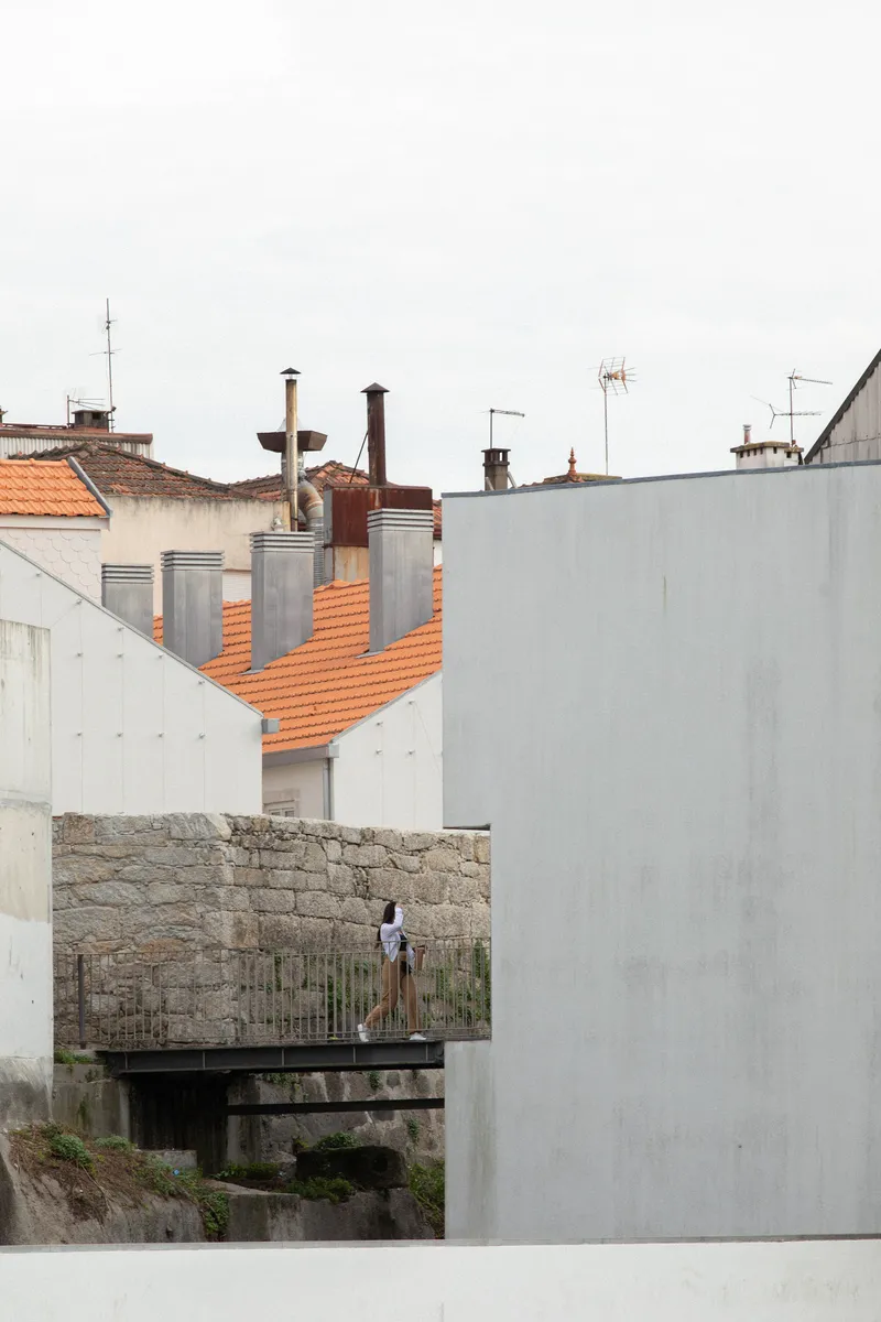 Figure standing on a timber footbridge between white concrete walls and terracotta rooftops
