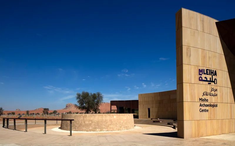Wood-clad entrance tower with signage beside a curved stone wall and desert landscape beyond