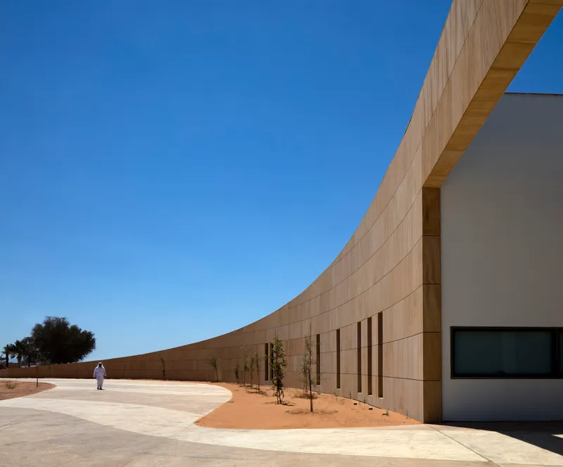 Curved sandstone wall with vertical window openings and a figure walking along the pathway