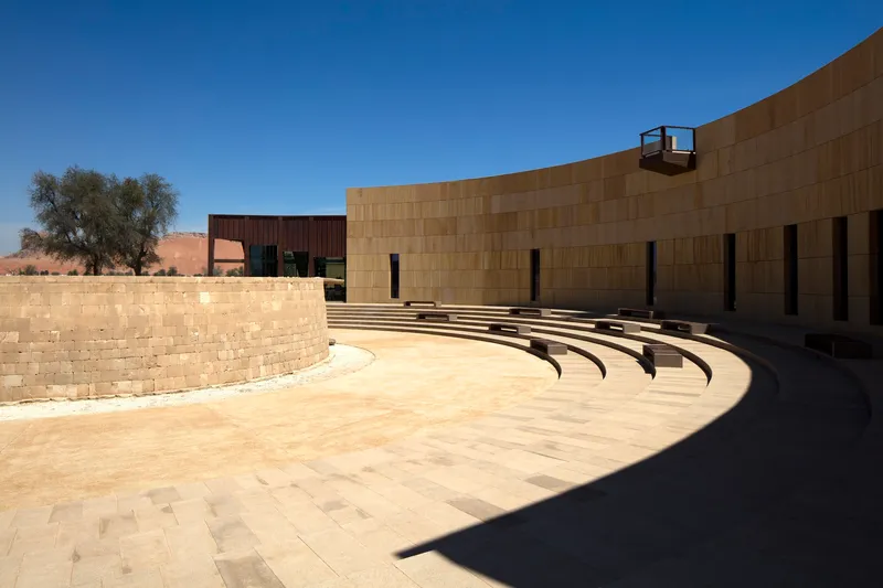Outdoor amphitheater with curved stone seating tiers and rammed earth walls under a clear blue sky