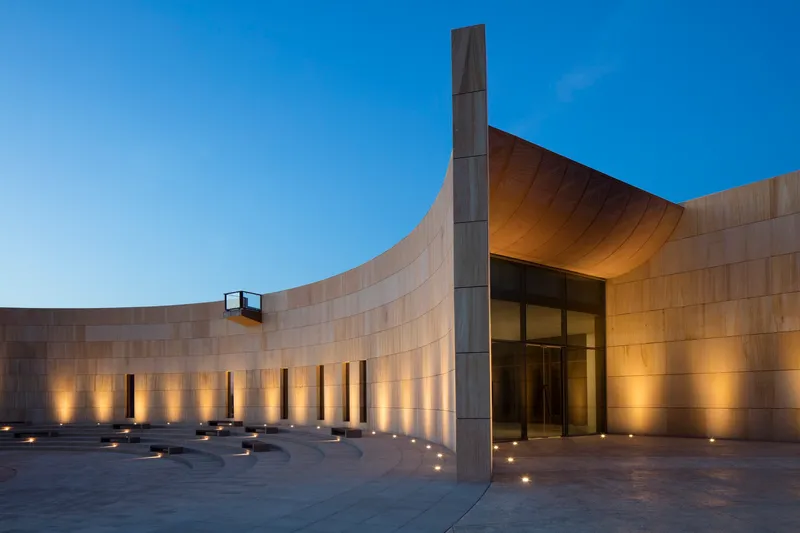 Curved rammed earth facade with uplighting and central entrance at dusk