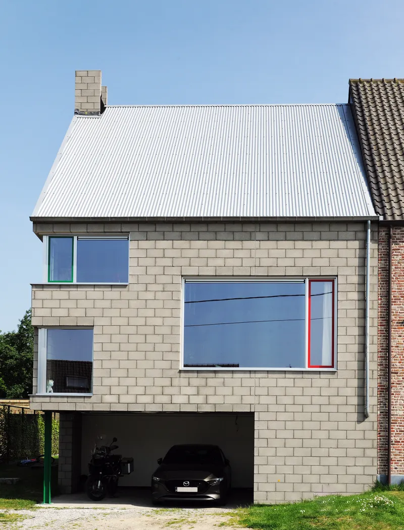 Front elevation of the pale masonry facade with corrugated metal roof and ground-level carport