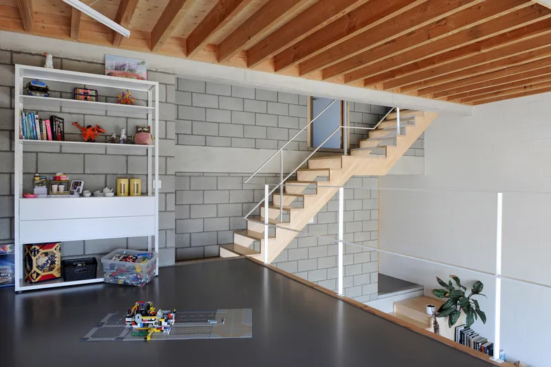 Interior view of floating timber staircase against concrete block wall with exposed rafter ceiling above