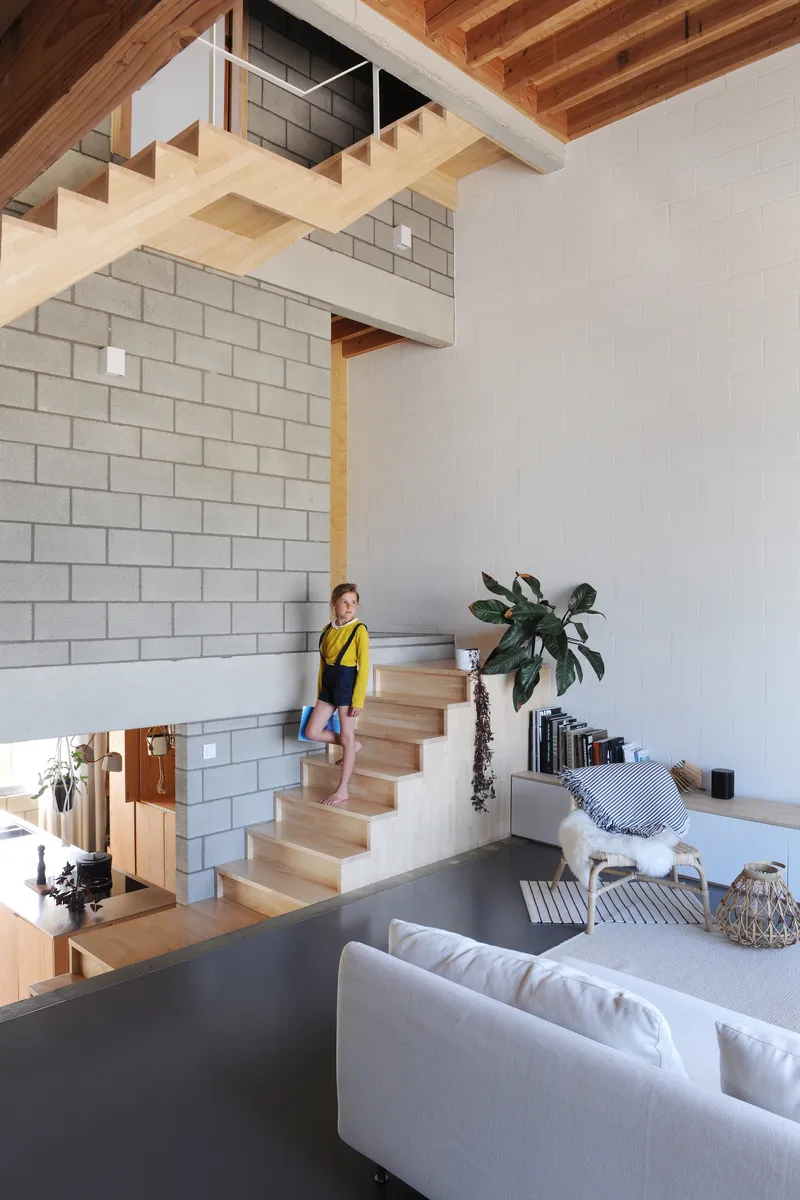 Double-height living space with open timber staircase and child ascending beside concrete block wall