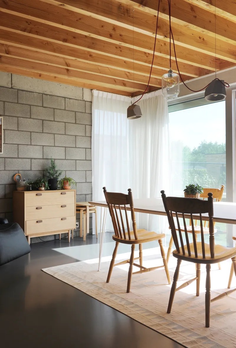 Dining area with timber ceiling joists and large corner window framing green landscape beyond white curtains