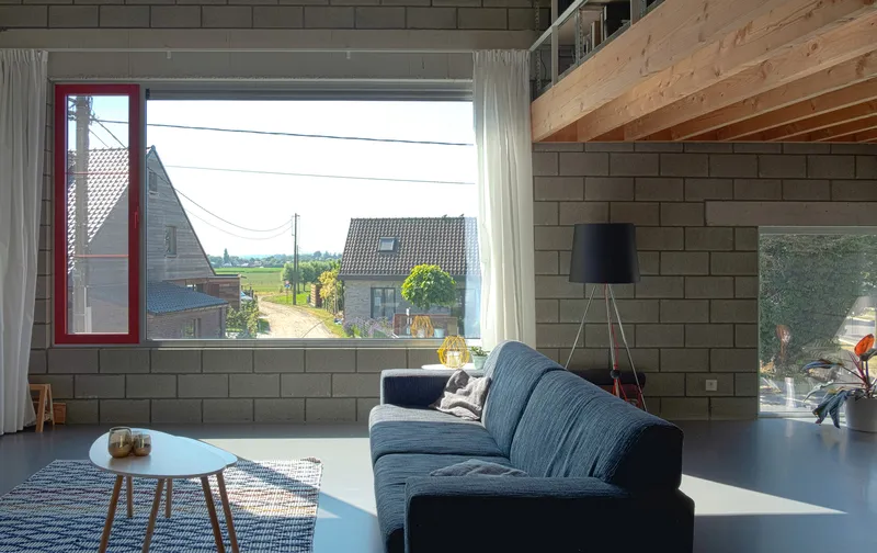 Living room with horizontal window framing neighboring rooftops and fields under exposed timber ceiling