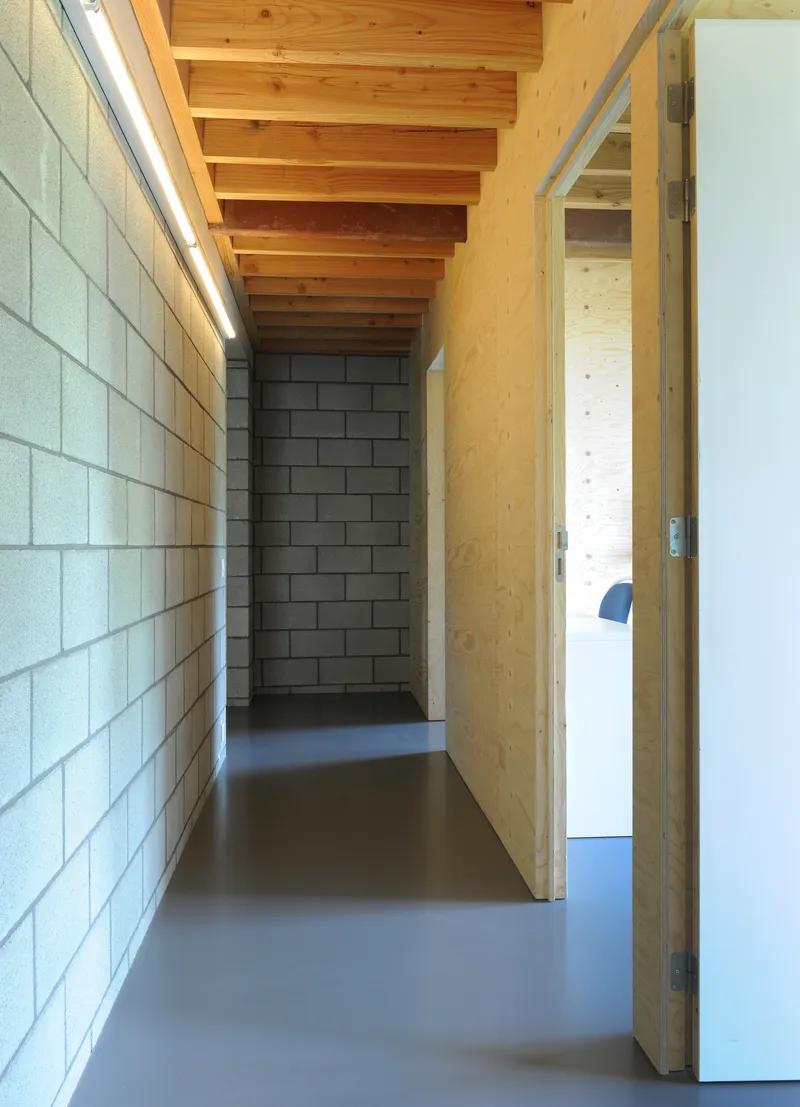 Narrow hallway with white concrete block walls, plywood partitions, exposed timber beams and polished concrete floor