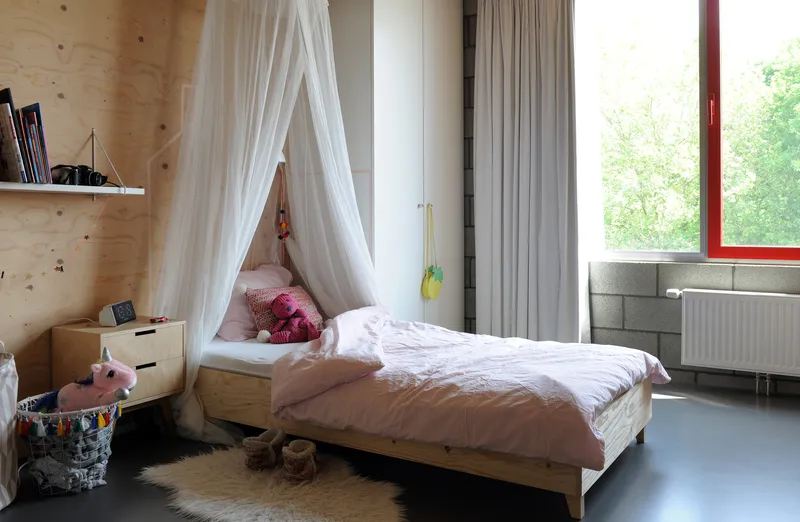 Children's bedroom with plywood walls, white canopy over a low bed, and red-framed window overlooking greenery