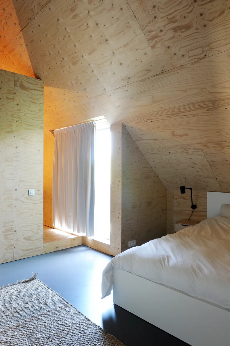 Bedroom with angled plywood ceiling and walls, bright sunlight streaming through curtained opening beside the bed