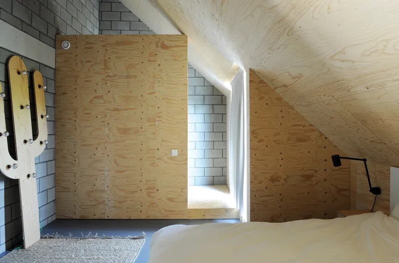 Corner bedroom view showing white concrete block wall, angled plywood surfaces and deep-set window framing daylight