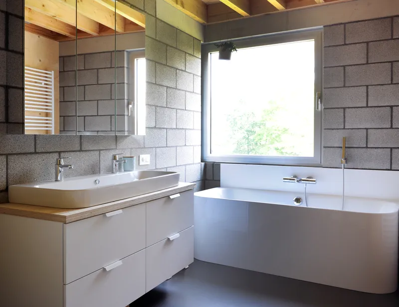 Bathroom with exposed concrete block walls, white vanity under timber-edged mirror, and freestanding tub below window