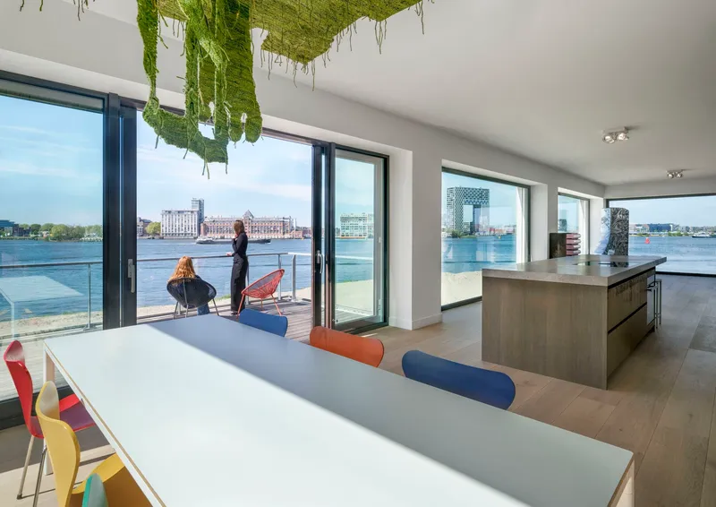 Open-plan living space with floor-to-ceiling glazing overlooking a waterfront and green textile installation overhead