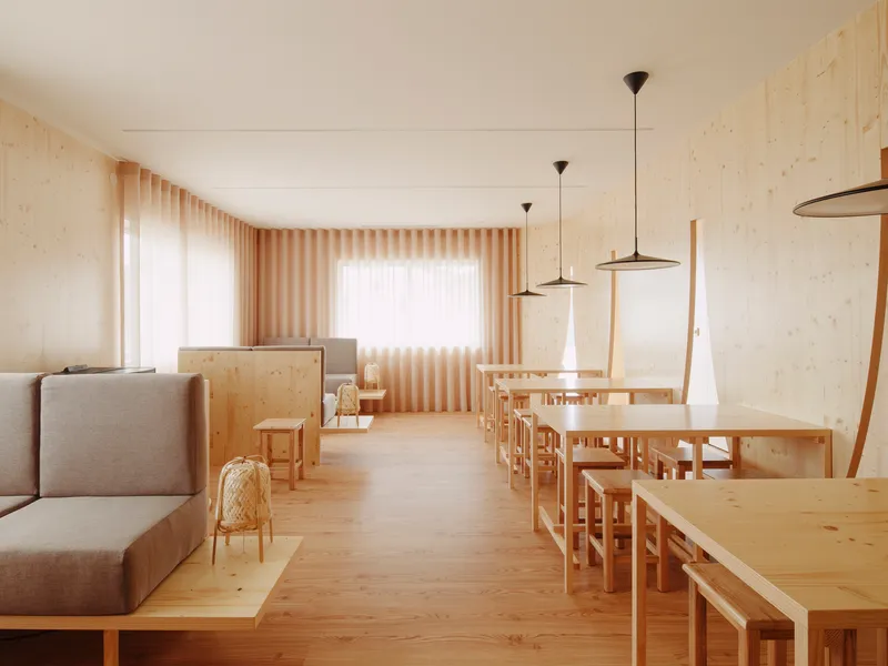 Dining hall with pale timber walls and rows of low tables with pendant lights filtering through sheer curtains
