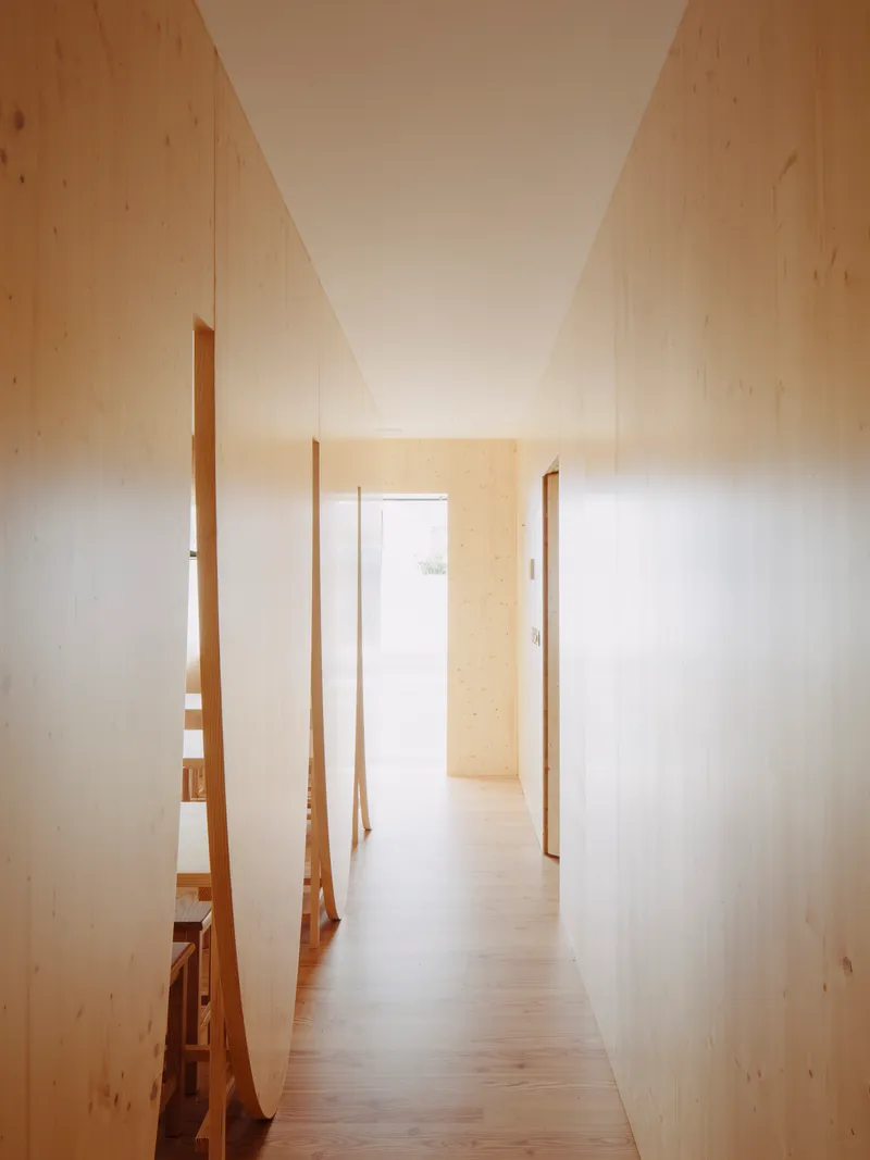 Narrow hallway with pale plywood walls and angled timber partitions opening to a sunlit doorway
