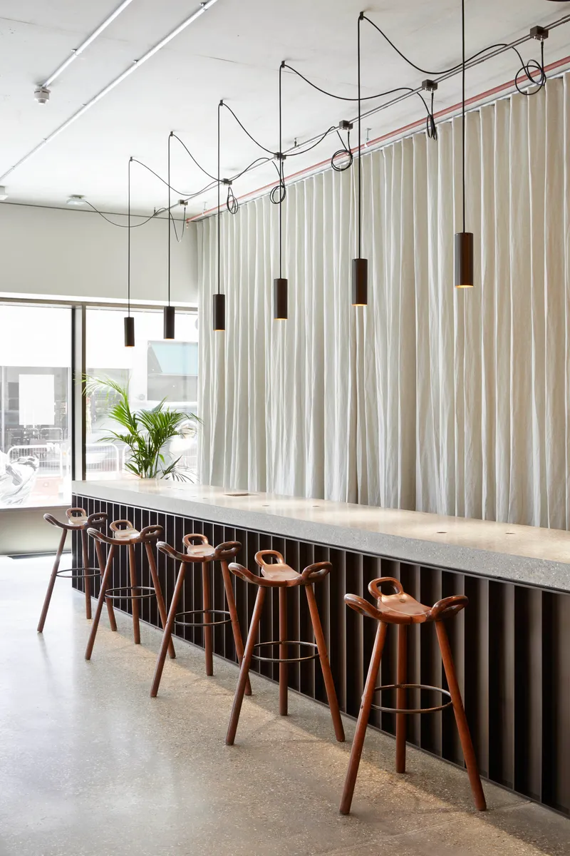 Long bar counter with timber stools below cylindrical pendant lights suspended from exposed conduit
