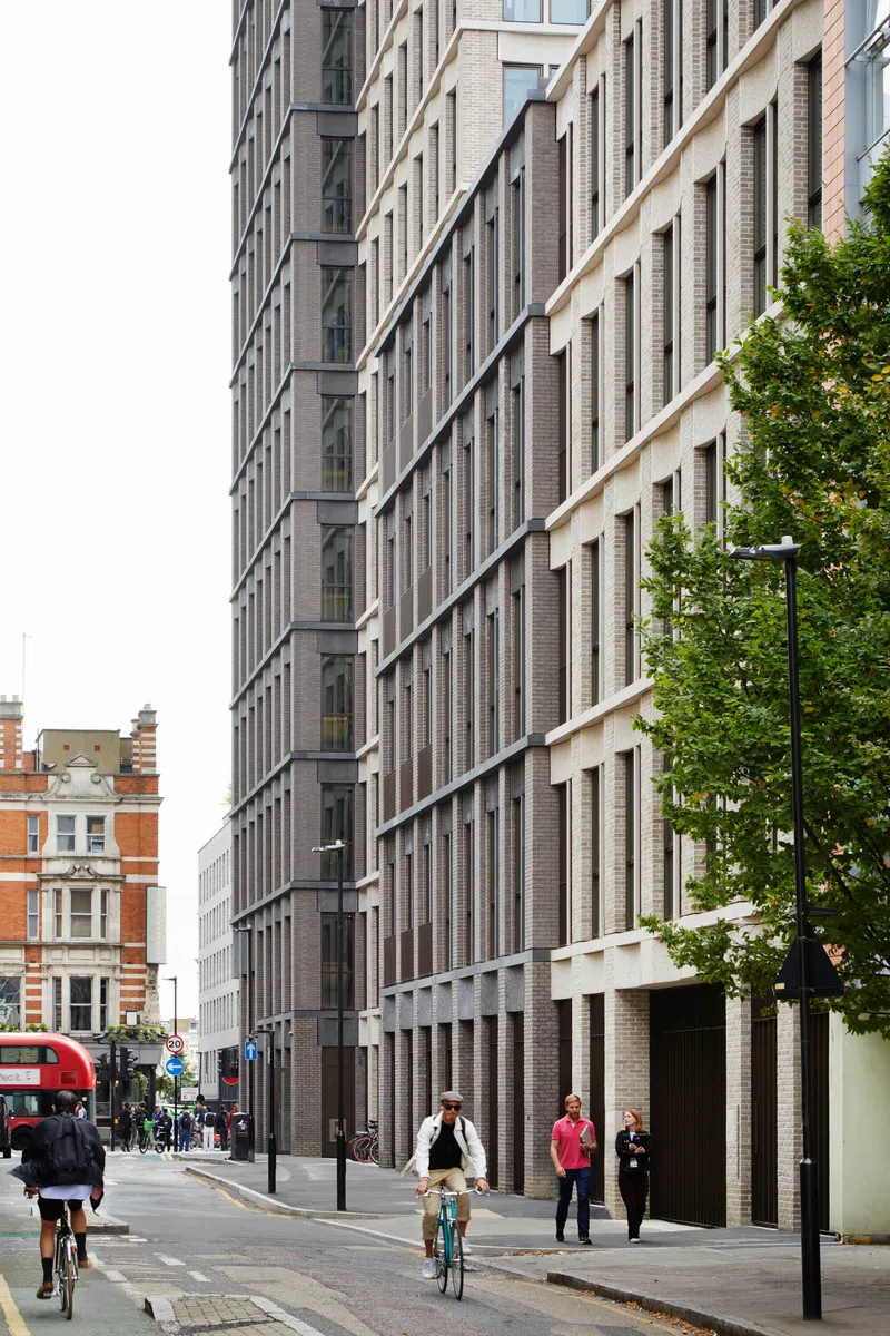 Street view of dark brick facade with vertical window recesses and cyclists passing below