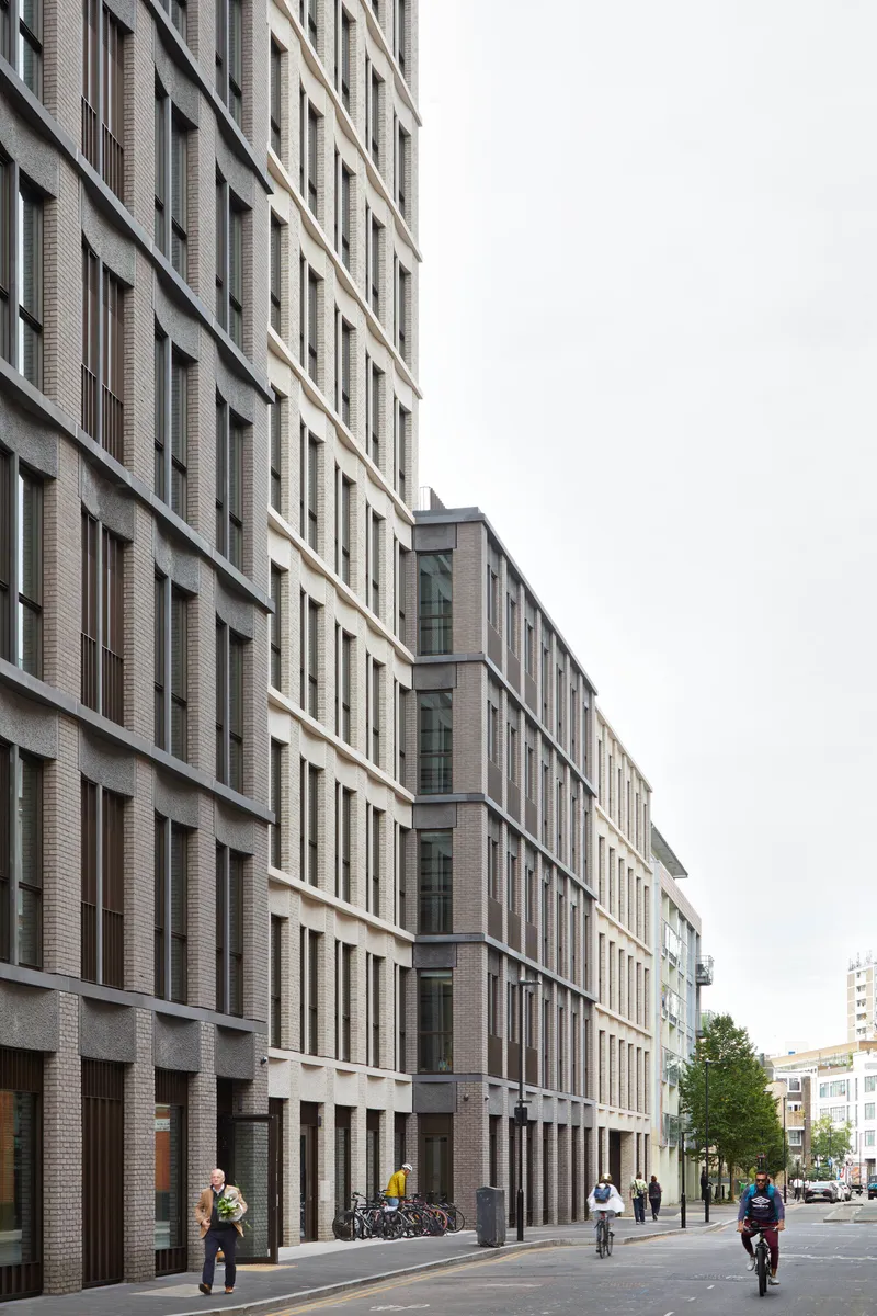 Grey brick facade along a street with pedestrians and cyclists under overcast sky