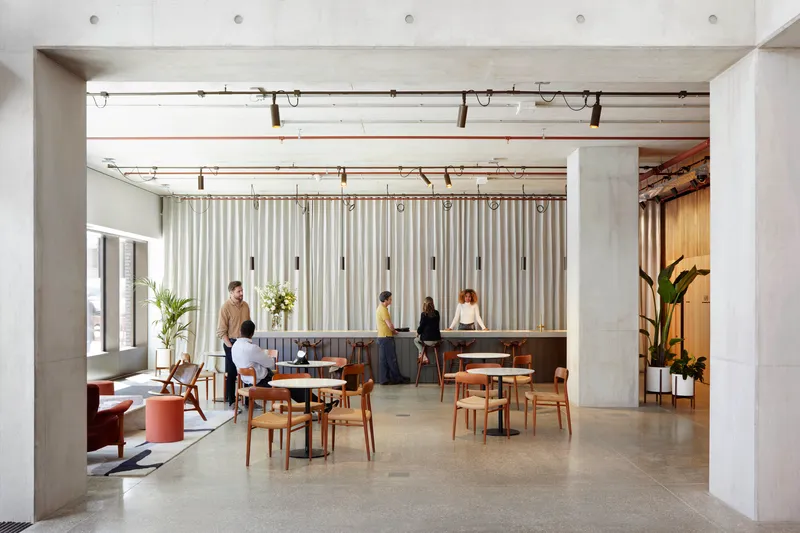 Open lounge area with exposed concrete beams, timber furniture and people gathering beneath pendant lighting