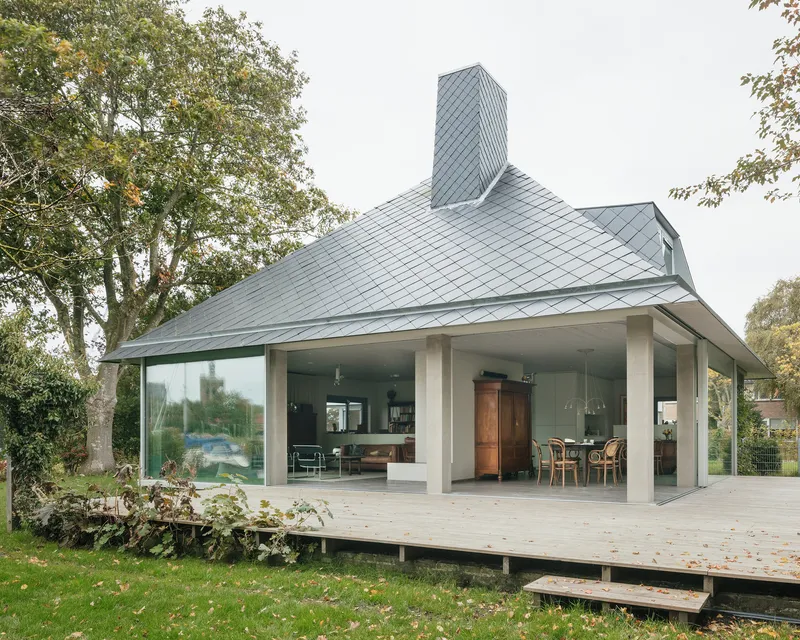 Covered terrace with white columns and floor-to-ceiling glass beneath a zinc-clad roof with chimney