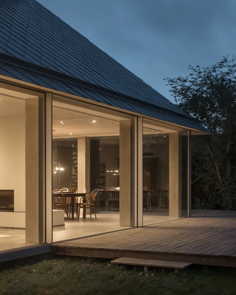 Sliding glass doors beneath the sloping metal roof opening to the timber terrace at dusk