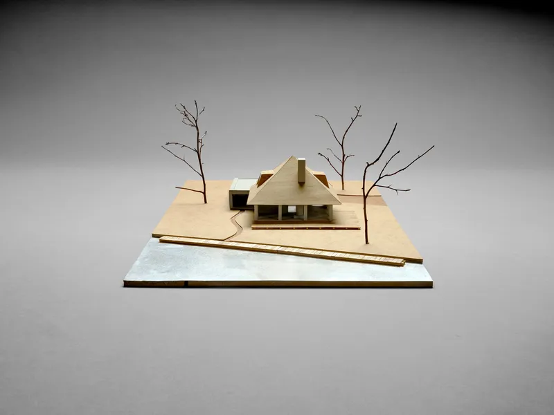 Wood physical model showing a gabled roof form with bare tree branches on the site