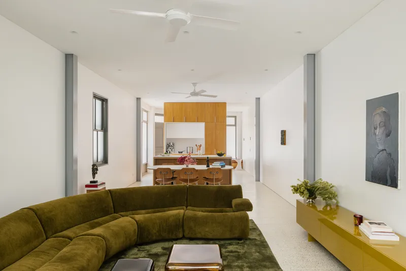 Living area with olive sectional sofa looking toward timber-clad kitchen volume beneath exposed steel columns