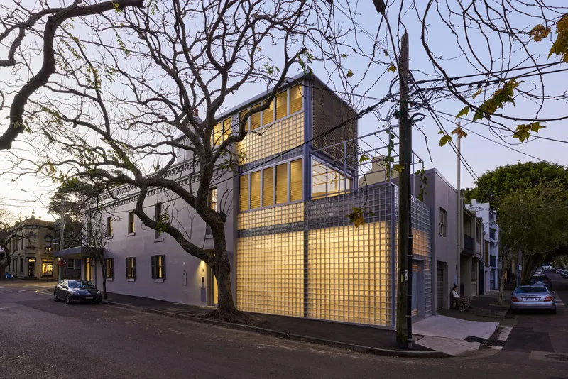 Oblique view from the street showing the illuminated glass brick volumes beside bare winter trees at twilight