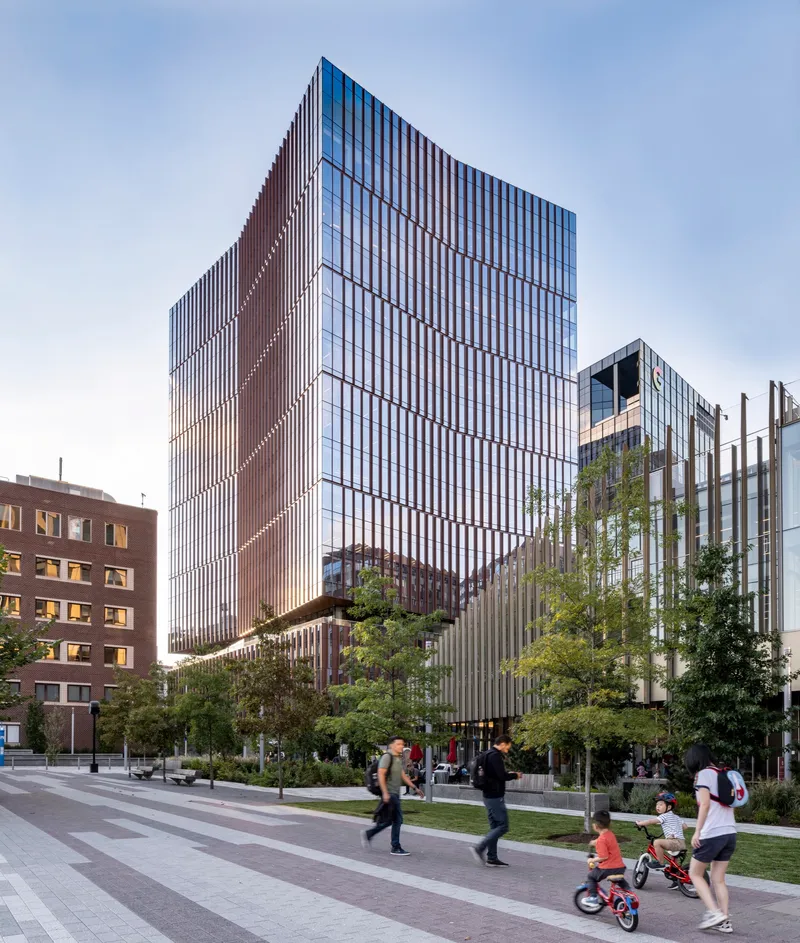 Vertical glass and fin facade with families on bicycles passing through the landscaped plaza at dusk