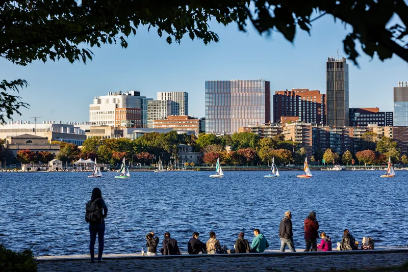 Skyline view from across the river with sailboats on the water and people gathered along the shore
