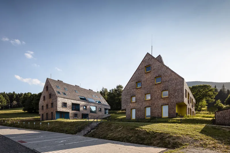 Two shingled gable-roofed volumes on a grassy slope with forests in the background
