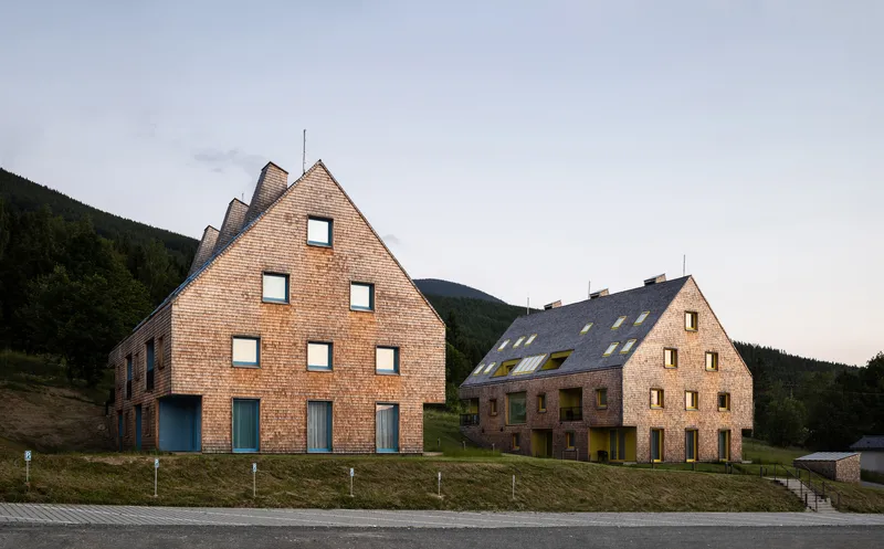 Street view of two timber-shingled gabled buildings set against wooded hills at dusk