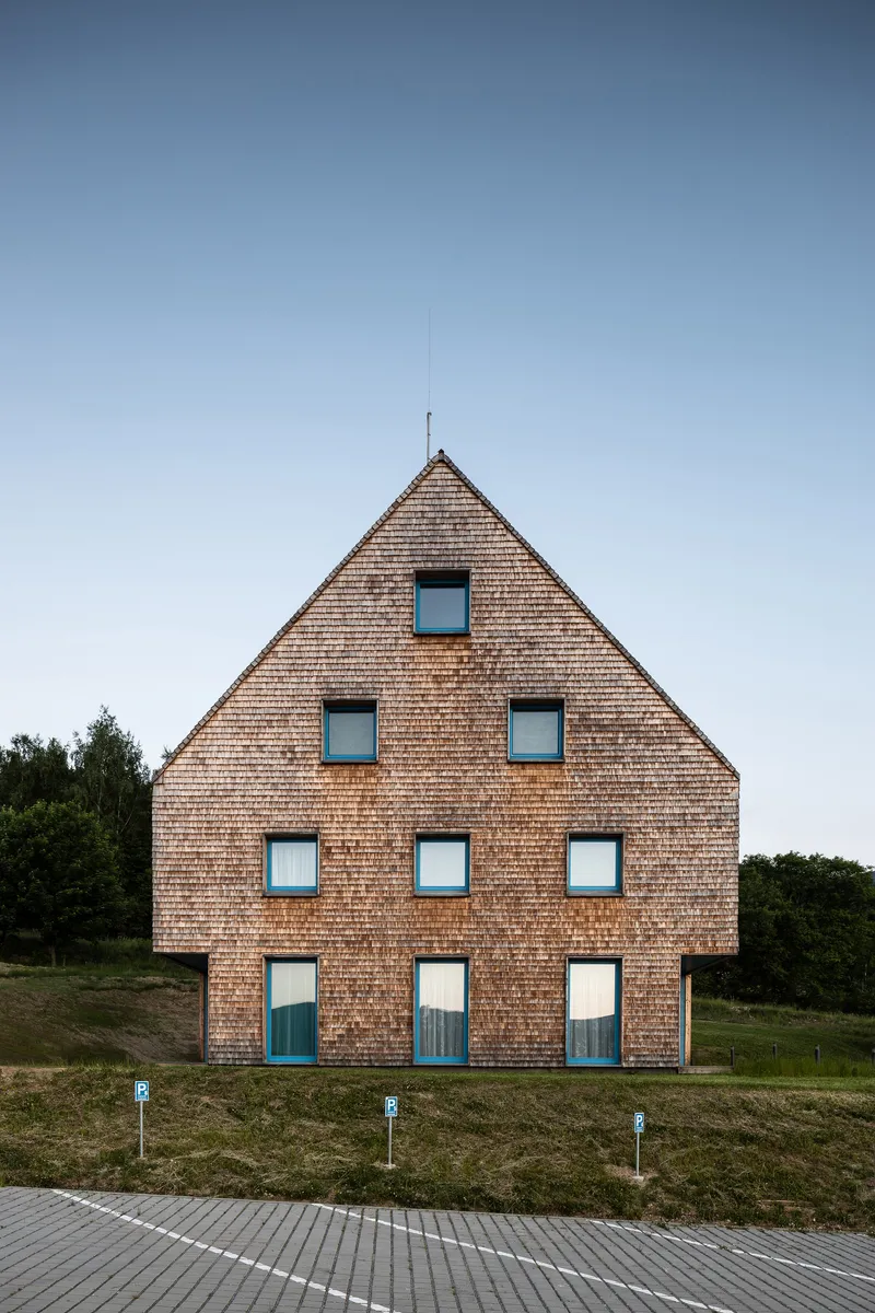 Front gable elevation with symmetrical square windows and teal doors on timber shingle cladding