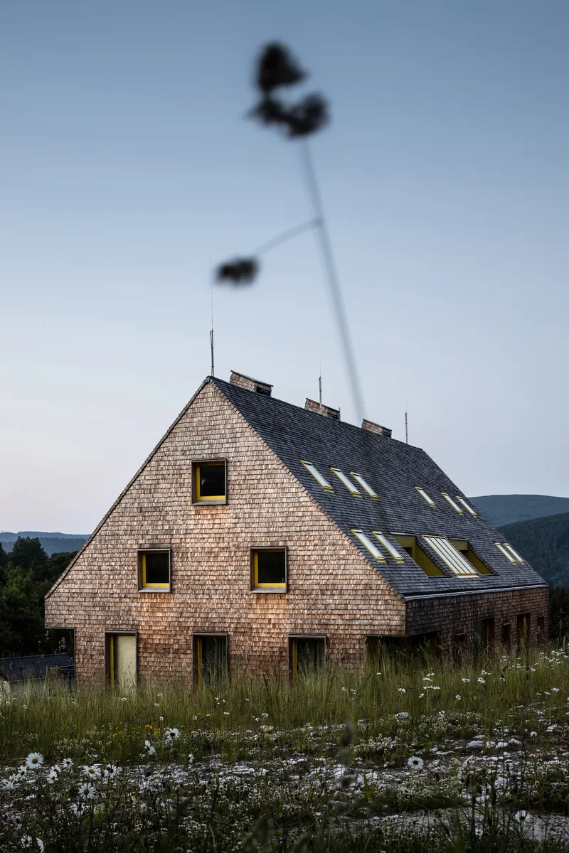 Gabled volume with timber shingle facade and skylight openings beneath a tall floodlight tower at dusk