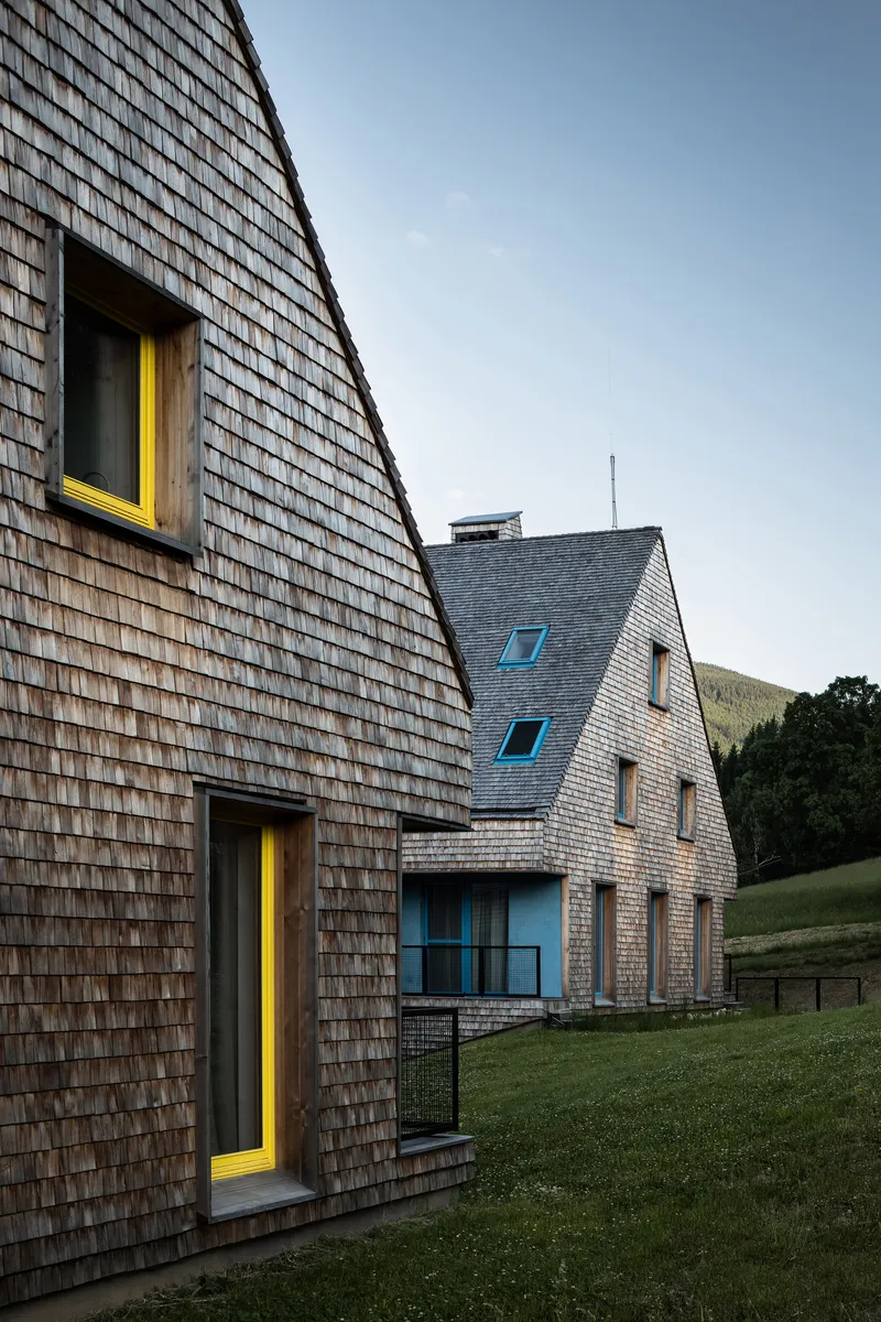 Steeply pitched shingled facade with yellow door frames at dusk on sloping grass terrain